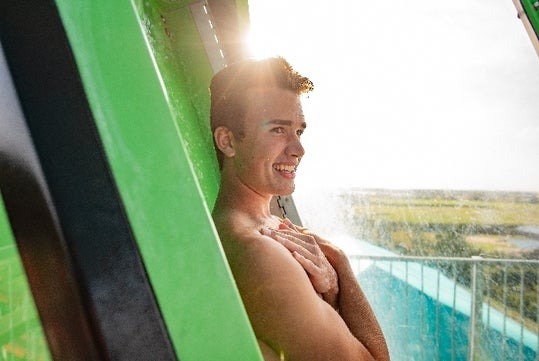 A young man stands inside a green water slide enclosure, smiling with hands crossed over his chest as sunlight shines behind him.