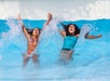 Two people in swimsuits raise their arms and splash water while playing in a bright blue swimming pool.