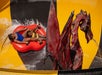 Two people ride a red inflatable float down a water slide, passing a large dragon statue that sprays water from its mouth.
