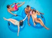 Four people relax on blue inflatable tubes in a pool, with two pairs holding hands and smiling at each other.