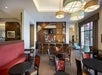 Modern hotel bar and dining area with round tables, red cushioned seating, wooden bar stools, large windows, and decorative light fixtures.