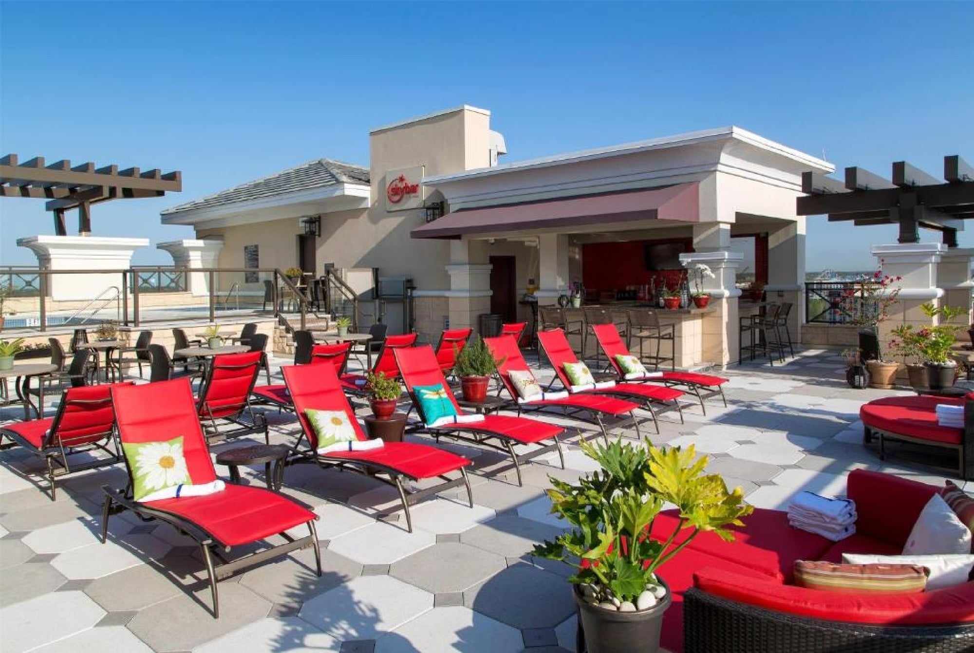 Outdoor rooftop lounge area with red cushioned chairs, small tables, decorative plants, and a bar under a clear blue sky.