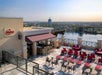 Outdoor rooftop bar with red lounge chairs and round tables overlooking a lake and cityscape under a clear sky during daylight.
