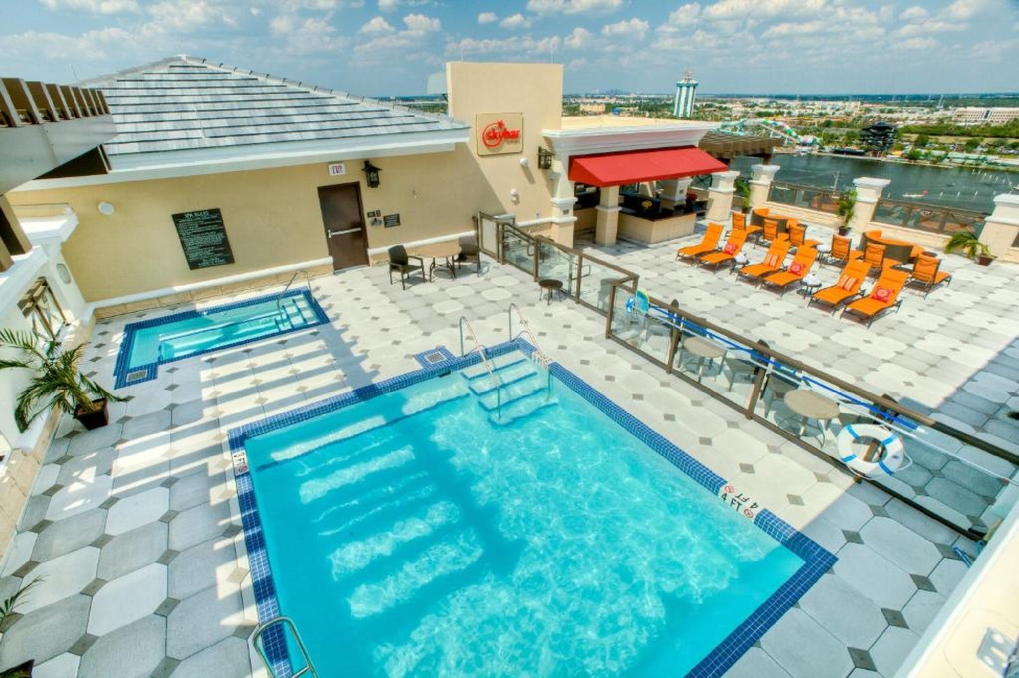 Rooftop pool area with a swimming pool, hot tub, orange lounge chairs, safety railings, and a small bar overlooking a cityscape under a partly cloudy sky.