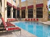 Outdoor swimming pool with clear water, surrounded by red cushioned lounge chairs and a beige building with large windows and red awnings.