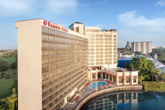 A large hotel building labeled "Ramada Plaza" with multiple floors, a pool area, and a waterfront view under a clear blue sky.