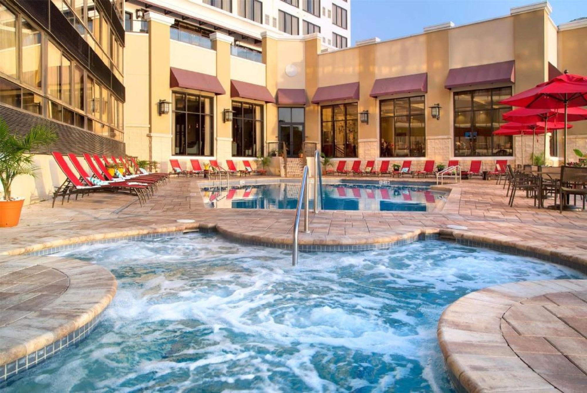 Outdoor hotel pool area with a circular hot tub in the foreground, lounge chairs, red umbrellas, and tables on a tiled patio, surrounded by tall buildings.