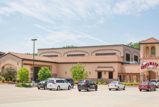 A Mediterranean-style restaurant with a parking lot in front, featuring several parked cars and greenery surrounding the building.