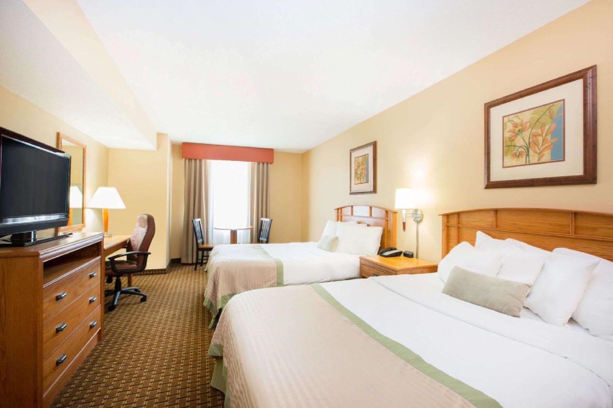 Hotel room with two double beds, a TV, wooden furniture, a desk with a chair, and a window with curtains. The walls are decorated with framed art.
