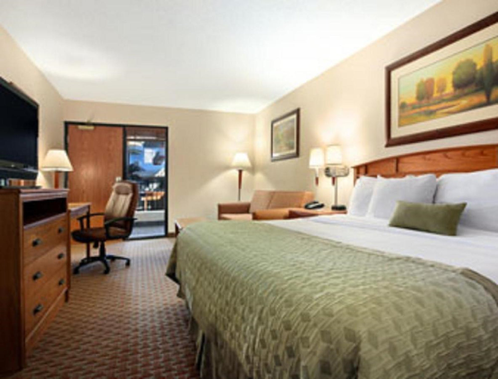 A hotel room with a large bed, green bedspread, and white pillows. There's a wooden dresser, TV, armchair, and a couch. Two framed paintings hang on the walls.