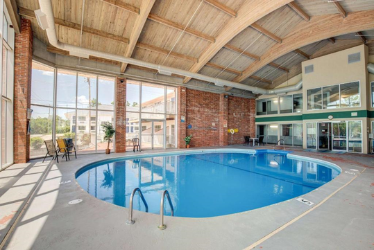 Indoor swimming pool with a curved roof, large windows, and brick walls. Tables and chairs are set up poolside.