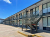 Exterior view of a two-story motel with outdoor staircases, large windows, and a parking area under a clear blue sky.