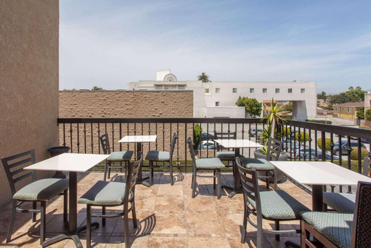 Outdoor seating area on a balcony with several tables and chairs, overlooking adjacent buildings and a partially visible parking lot under a clear sky.