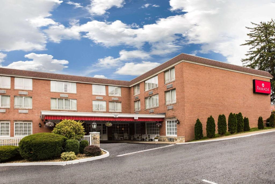 Brick hotel building with red awning, multiple windows, and a Ramada sign. Surrounded by shrubs and a sloped driveway. Cloudy sky in the background.