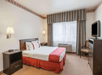 Hotel room with a double bed, red-and-white bedding, nightstand, lamp, striped curtains, wall-mounted TV, small dresser, and a chair under a window. Beige carpet and patterned border ceiling.