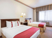 Hotel room with two double beds, each featuring red runners and white pillows. A nightstand with a phone and lamp is between the beds. A window with striped curtains is on the right wall.