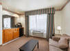 Hotel room with a sitting area, featuring a couch, coffee table, lamp, kitchenette with a sink, microwave, and coffeemaker. There's a large window with striped curtains and an air conditioner.