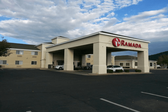 Exterior view of a Ramada hotel with beige facade, covered entrance, and parked cars in front under a partly cloudy sky.