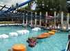 People at a water park cross a pool by stepping on floating platforms shaped like fruit slices and clouds, with a net overhead for support.