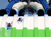 Two people on blue mats slide down parallel water slides with blue, green, and white lanes, splashing water as they descend.