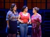 Three women stand on stage under warm lighting, holding hands and facing each other, with shelves and large windows in the background.