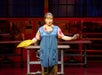 A performer in costume stands on stage holding a yellow fan, with arms partially extended, in a room resembling a workshop or sewing studio.