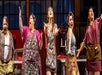 Five women on stage passionately singing, wearing colorful aprons. Backdrop includes sewing machines and fabric, indicating a musical or theatrical performance setting.