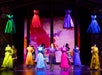 Stage performance featuring actors surrounded by colorful dresses on mannequins and suspended above, set against a bright, floral backdrop.