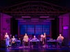 Six actors stand and sit at sewing tables on a brightly lit stage set resembling a garment workshop, with large windows and sewing equipment visible in the background.