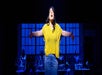 Person on stage wearing a yellow top and jeans, arms spread wide, singing against a backdrop of blue-lit window panes with people working behind.