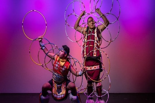 Two performers in traditional attire are holding and surrounded by multiple hoops. The background is lit with a purple hue.