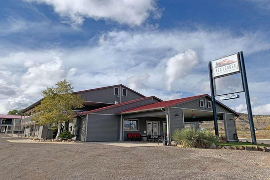 A roadside motel with a red roof and a sign reading "Red Ledges." The building is surrounded by a gravel parking area and sparse vegetation, with a partly cloudy sky overhead.