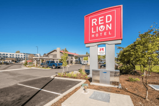 The red sign for the Red Lion Hotel in their parking lot with the hotel in the background on a sunny day in Portland.