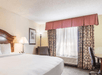 A hotel room with a king bed, an accent chair and lamp, a desk and office chair, and a large window with curtains on the far wall.
