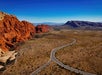 A winding road passes through a desert landscape with red rock formations on the left and distant mountains under a clear blue sky.
