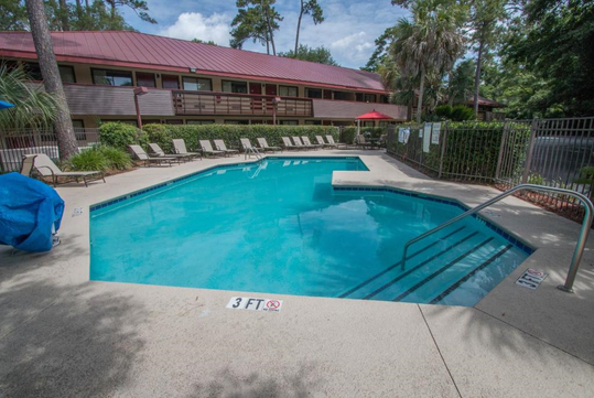 Outdoor pool with lounge chairs and a surrounding fence, located in front of a building with red roofing. Lush green trees are visible in the background.