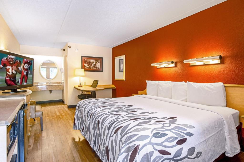 A hotel room with a large bed, patterned bedspread, wall-mounted TV showing a sports game, desk with a laptop, chair, and a lit vanity mirror next to the washbasin.
