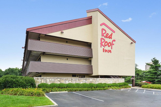Three-story Red Roof Inn building with beige walls, red roof accents, and a visible sign. The foreground shows a parking lot and landscaped greenery.