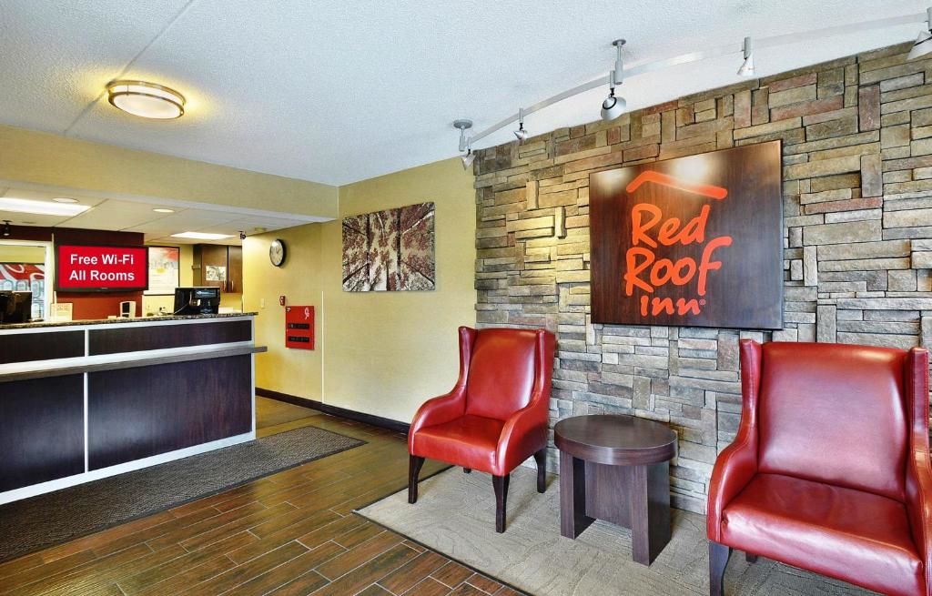 Hotel lobby with two red chairs, a small table, and a Red Roof Inn sign on a stone accent wall. Front desk area on the left with a "Free Wi-Fi All Rooms" sign.
