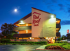 Night view of a three-story Red Roof Inn with illuminated signage. The building is surrounded by greenery and a clear sky with a visible moon.