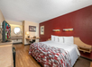 Hotel room with a king-sized bed, red accent wall, and wooden flooring. Includes a flat-screen TV, desk, and a vanity area with a round mirror in the background.