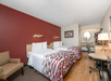 Hotel room with two double beds, patterned bedspreads, a side table, chair, desk with a lamp, and a wall-mounted TV. Red accent wall behind the beds, round mirror near the sink area.