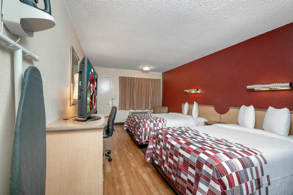 Hotel room with two beds featuring checkered bedding, a wall-mounted TV, desk, chair, ironing board, and red accent wall.