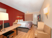 Hotel room with a double bed, red accent wall, desk with lamp, TV, and armchair on wooden flooring.