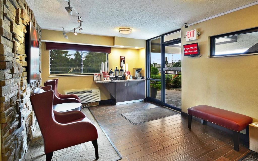 Hotel lobby with red chairs, a bench, a coffee station on a counter, and a door leading outside. Brown tiles cover the floor, and a stone accent wall is on the left.
