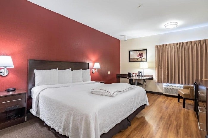 Hotel room with a large bed, white bedding, red accent wall, two bedside lamps, a desk, chair, window with curtains, wall art, and wooden floor.