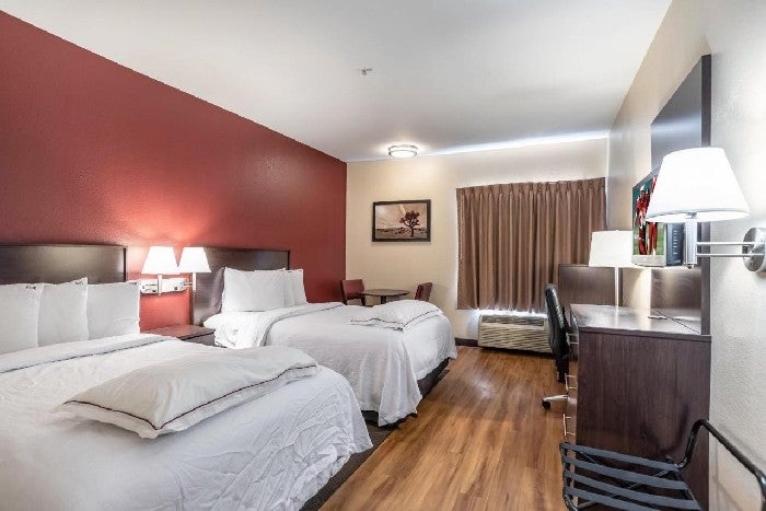 Hotel room with two double beds, a desk with a chair, a wall-mounted lamp, a small table with two chairs, and a window with closed curtains.