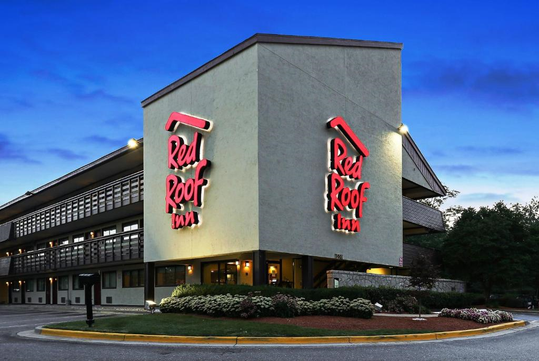 Exterior view of a Red Roof Inn at dusk, featuring red signage on the corner of the building, with a parking area and landscaped bushes.