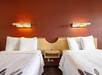 Two beds with white linens and pillows are separated by a small wooden nightstand, beneath two modern wall lights on a red wall.