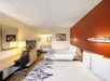 Hotel room with two double beds, wall-mounted lights, a desk with chair, a TV, and a vanity area with a mirror in the background.